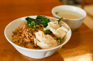 A bowl of Indonesian chicken noodles (Yamie) with wontons and a side of broth on a wooden table.