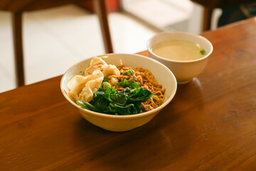A bowl of Indonesian chicken noodles (Yamie) with wontons and a side of broth on a wooden table.