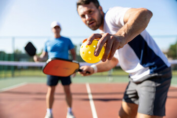 A dynamic sports photograph captures the precise moment a pickleball player prepares to serve on a sun-drenched outdoor court.