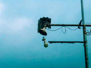 A broken street lamp hangs loosely from a rusty pole under a cloudy sky.