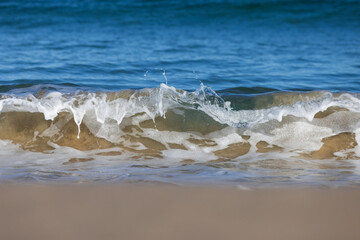 A small shorebreak wave crashes along a sandy beach creating abstract shapes and textures on a...