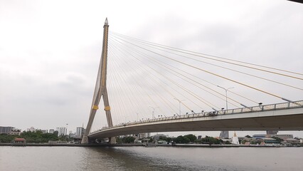 Large cable-stayed bridge crossing a wide river with tall central pylon and golden suspension cables, urban skyline and riverside buildings visible in the background under cloudy sky.