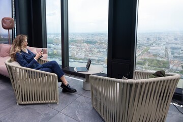 In a stunningly designed lounge area, a stylish woman is seated comfortably, unwinding while taking in the breathtaking panoramic views of the vibrant city skyline through a tall highrise window