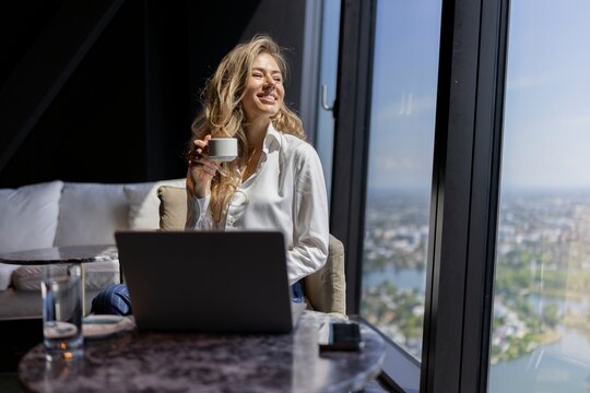 A joyful woman happily sipping coffee while working on her laptop in a stylish, modern highrise apartment, showcasing a breathtaking city view that inspires creativity and comfort