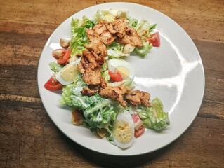 Caesar salad with chicken. With a mix of eggs, tomatoes, lettuce, onions, Parmesan cheese, croutons, and Caesar dressing. A wooden table background.