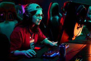 Caucasian young adult woman wearing headset sitting at gaming desk playing computer game in esports...