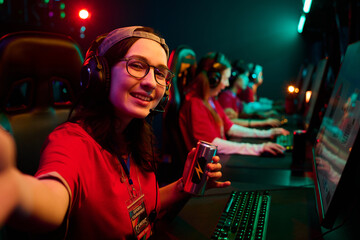 Portrait of Caucasian teenage girl wearing headset smiling and taking selfie while holding energy drink can, sitting at gaming desk with multiethnic teenage girls playing video games in esports team © pressmaster