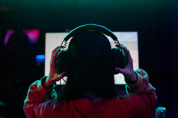 Teenage girl with long dark hair wearing headphones sitting in front of computer monitor gaming in...