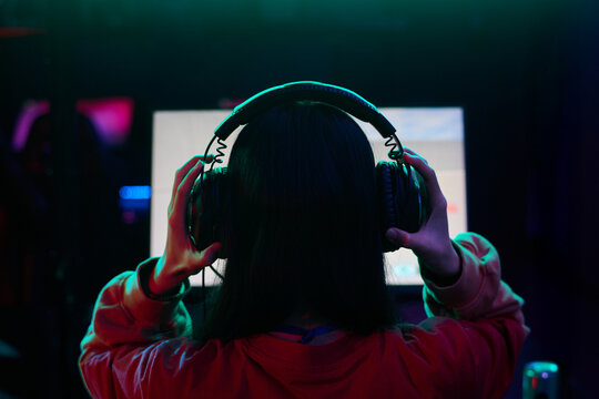 Teenage girl with long dark hair wearing headphones sitting in front of computer monitor gaming in dimly lit room - Powered by Adobe