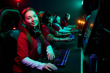 Teenage girl gamer sitting at computer desk wearing headset looking at camera with multiethnic...