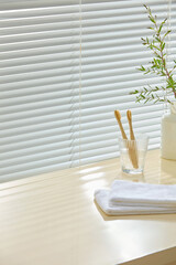 Bathroom setup with soft tile wall, towels, plant vase and toothbrushes. Light wood surface left...