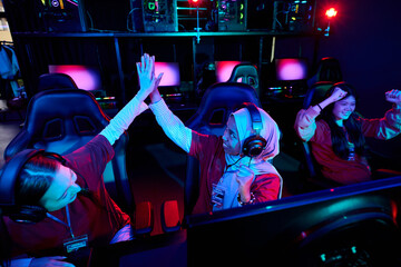 Diverse team of young adult and teenager Asian and Caucasian women wearing headsets celebrating esports victory in gaming arena, sitting at gaming desks with monitors and gaming chairs