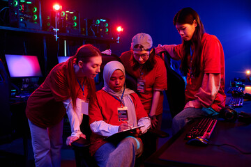 Group of multiethnic teenage girls gamers discussing strategy around gaming chair in esports arena,...
