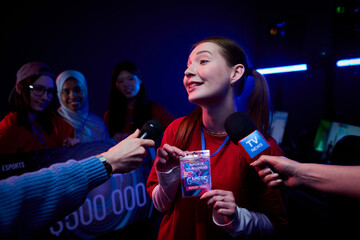 Caucasian young adult woman holding e-sports tournament badge and speaking to reporters with microphones while diverse team of teenage girls gamers