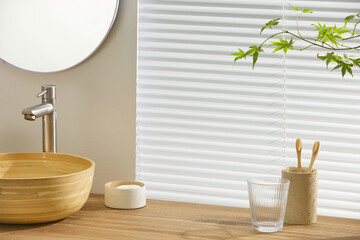 Glass cup with toothbrushes and warm wood sink against clean blinds. Aesthetic balance of texture...