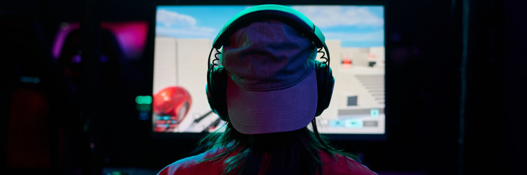Teenage girl wearing headphones sitting in front of large gaming monitor playing video game in dark room, back view showing focused posture, participating in girls gamers team event