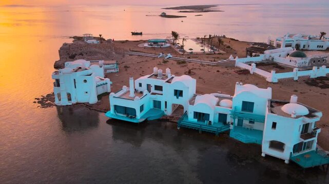 Several white houses floating on the water with rock jetties and blue houseboats. The sky is orange and pink, creating a beautiful sunset atmosphere over the lake. Location: Tunisia, Medenine ,Zarzis