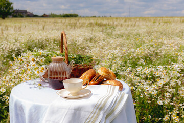 A table set for tea with pastries, a teapot with tea, a cup and a bouquet of daisies in a basket,...