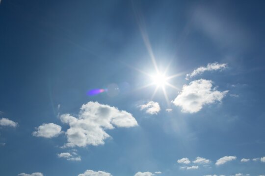 Naklejki Flat design showing bright sun casting starburst lens flare over deep blue sky with fluffy clouds