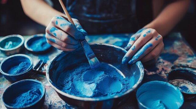 Close-up of hands mixing blue pigment powder with brush in a bowl, surrounded by containers of blue paint. - Powered by Adobe
