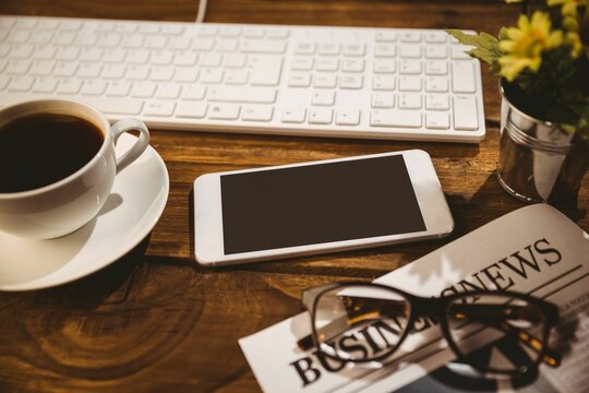 Flat design wooden desk is featuring centered smartphone, keyboard, coffee cup, newspaper, flowers