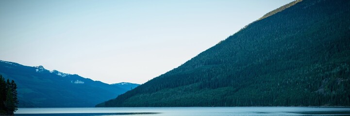 Depicting flat design lake reflecting steep forested mountain slope and snow peaks under clear sky