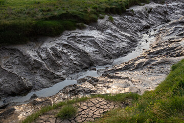 Muddy channel with exposed banks and a small stream of water, bordered by grassy slopes and cracked earth