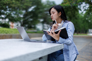 Young asian woman lies outdoors in park reading book using laptop computer Education studying...