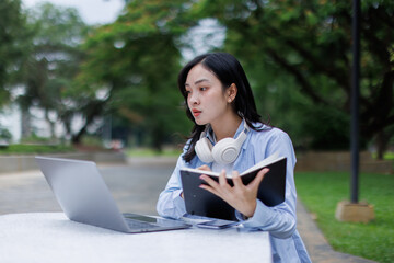 Young asian woman lies outdoors in park reading book using laptop computer Education studying...