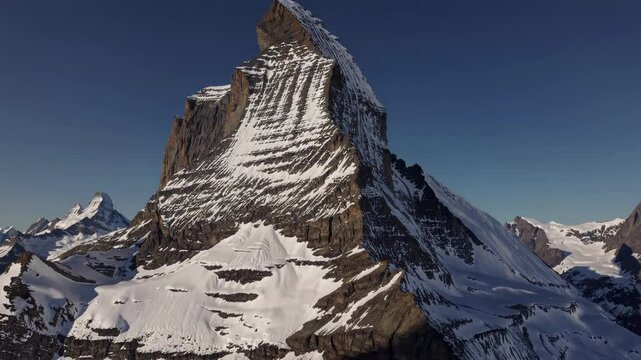 Aerial video captures a majestic snow-capped mountain peak under a clear blue sky, showcasing its rugged texture and towering presence from a high angle.