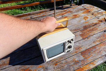 A portable retro television with a retractable antenna stands on a wooden table outdoors. A hand...