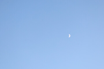 A young moon is clearly visible against a cloudless, pale blue daytime sky. Its delicate crescent...