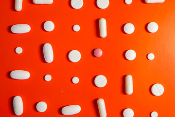 White pills and capsules, along with one pink one, are evenly laid out on a bright orange background. This visually appealing composition symbolizes the pharmaceutical industry and the variety of medi