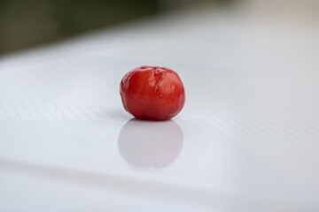 A single ripe red acerola cherry on a clean white reflective surface. A minimalist still life photo representing simplicity, freshness, and healthy eating.

