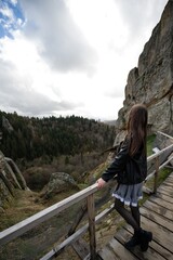 Girl gazing at a scenic valley view from a wooden bridge overlooking rocky terrain and forest.