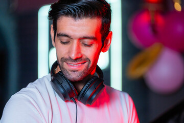 A handsome DJ focuses on his music set in a vibrant nightclub with a guest in the background A...