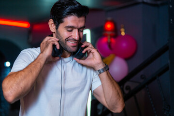 A handsome DJ focuses on his music set in a vibrant nightclub with a guest in the background A...