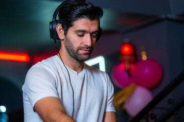 A handsome DJ focuses on his music set in a vibrant nightclub with a guest in the background A...