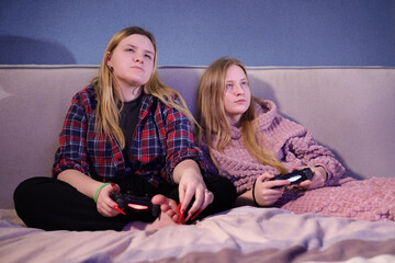 Two teen girls playing video games on a bed, both intensely focused on the screen, holding wireless...