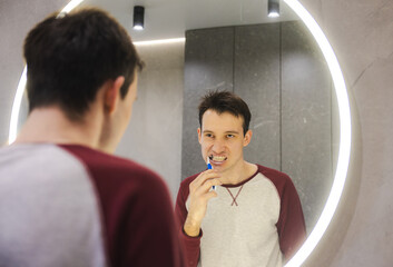 Cheerful young man brushes his teeth in front of a bathroom mirror, focusing on cleanliness and...