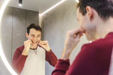 Young man flossing his teeth in front of the mirror, smiling slightly as he maintains good oral...