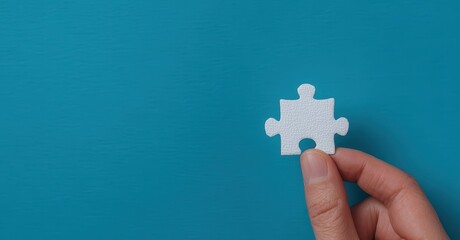 Obraz premium A hand holding a single white puzzle piece against a blue background symbolizes problem-solving and fitting pieces together.