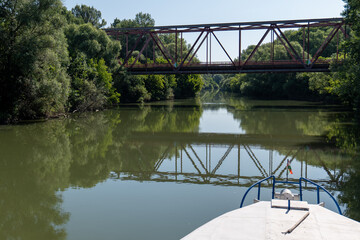 Boat with Hungarian flag on Bodrog river passing under metal bridge in Viničky, Slovakia. Calm water reflects the bridge and green trees on a sunny summer day.