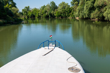 Boat on Bodrog river with Hungarian flag, green trees, calm water, blue sky and mooring rope on the deck. Summer river landscape between Slovakia and Hungary.