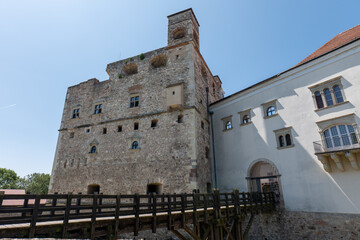 Stone castle tower and white building with wooden bridge in Sarospatak, Hungary, under clear blue sky.