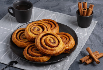 Homemade freshly baked cinnamon buns in black plate with cinnamon sticks, coffee and napkin on grey table