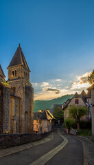Winding cobblestone street through the historic French village of Conques at golden hour with the evening light illuminating the tower of the Abbey of Sainte-Foy and medieval houses under a blue sky,