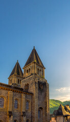 The twin spires of the ancient towers of Sainte-Foy Abbey and medieval houses are illuminated by the golden light of sunset under a clear blue sky in Conques, France.