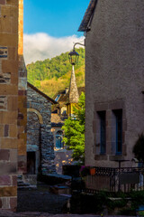 A hidden alleyway off a medieval street with the golden light of sunset illuminating the spire of a stone church tower and the green hills in the background in Conques, France.