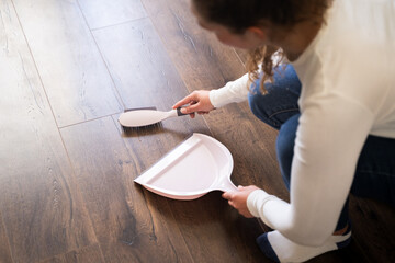 Woman sweeping wooden floor with pink small plastic dustpan and brush indoors, doing household chores, home cleaning routine, cleanup housework, top view of hands and tools on laminate surface